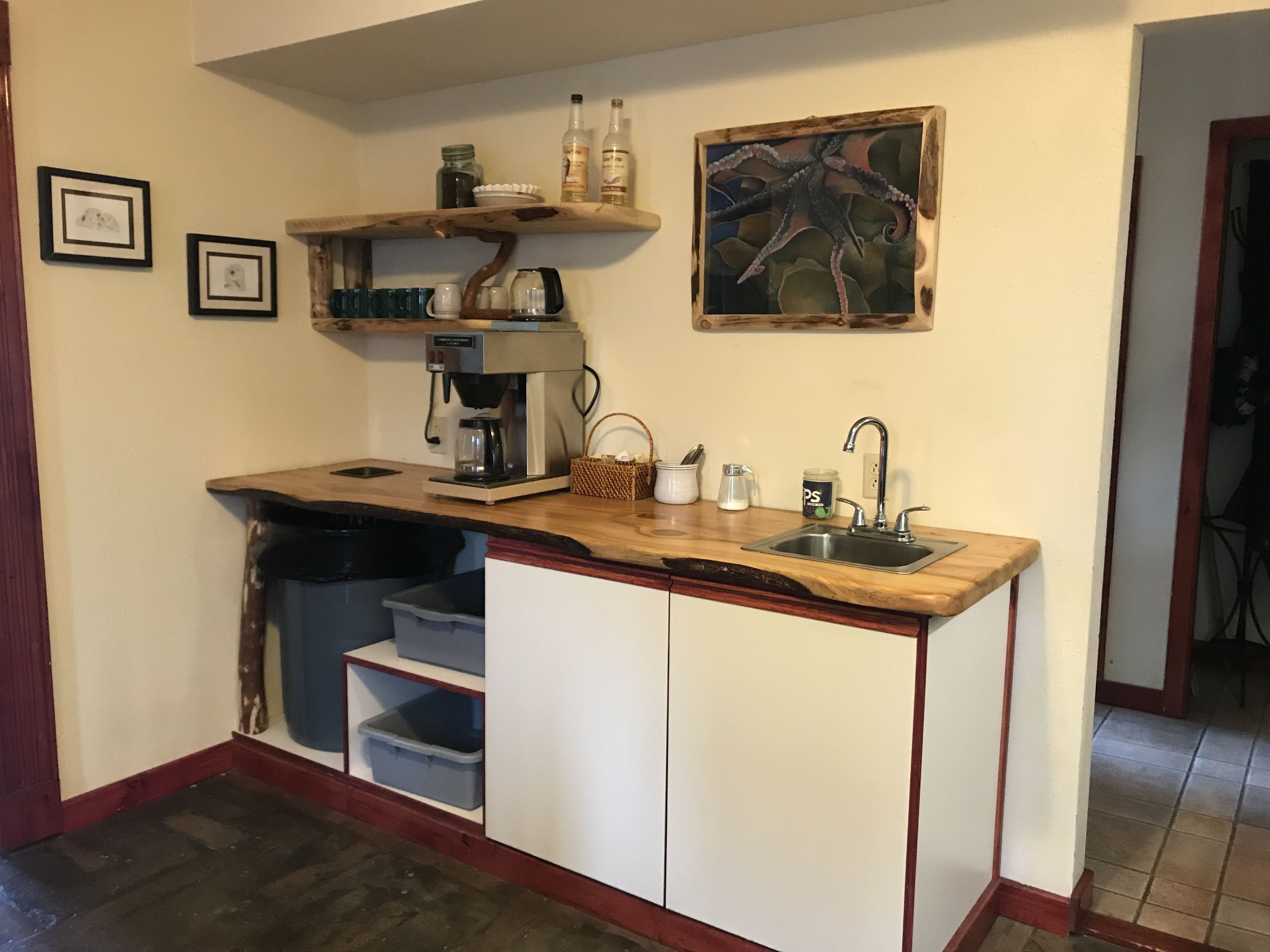 Cocina compacta con mostrador de madera, cafetera, lavamanos y estante con diversos objetos.