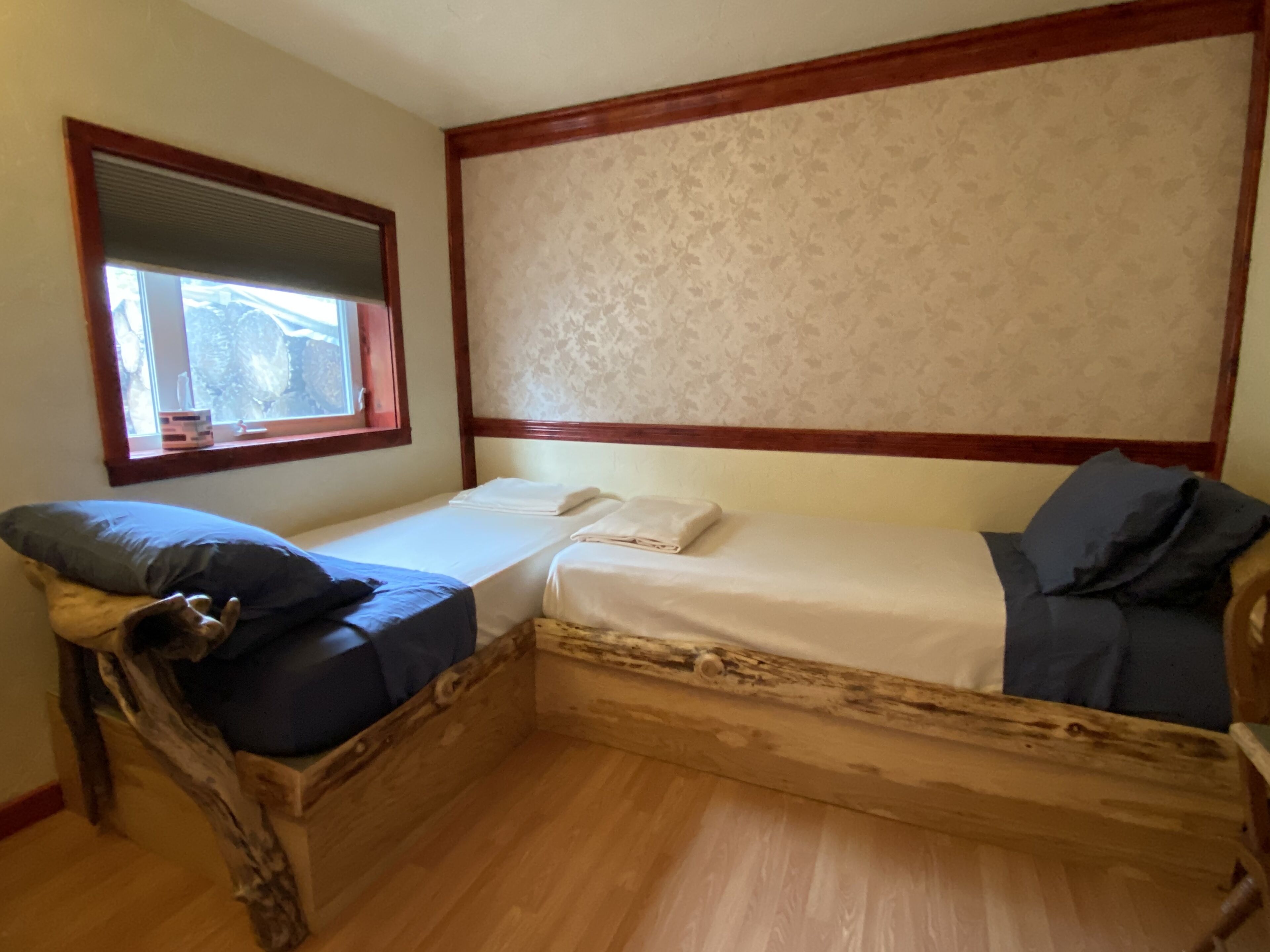 A small, single-bed room with a wooden headboard, a window with blinds, and a patterned wall.