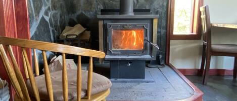 A stone wall, a wooden chair, a fireplace with a visible fire, and a window with daylight.