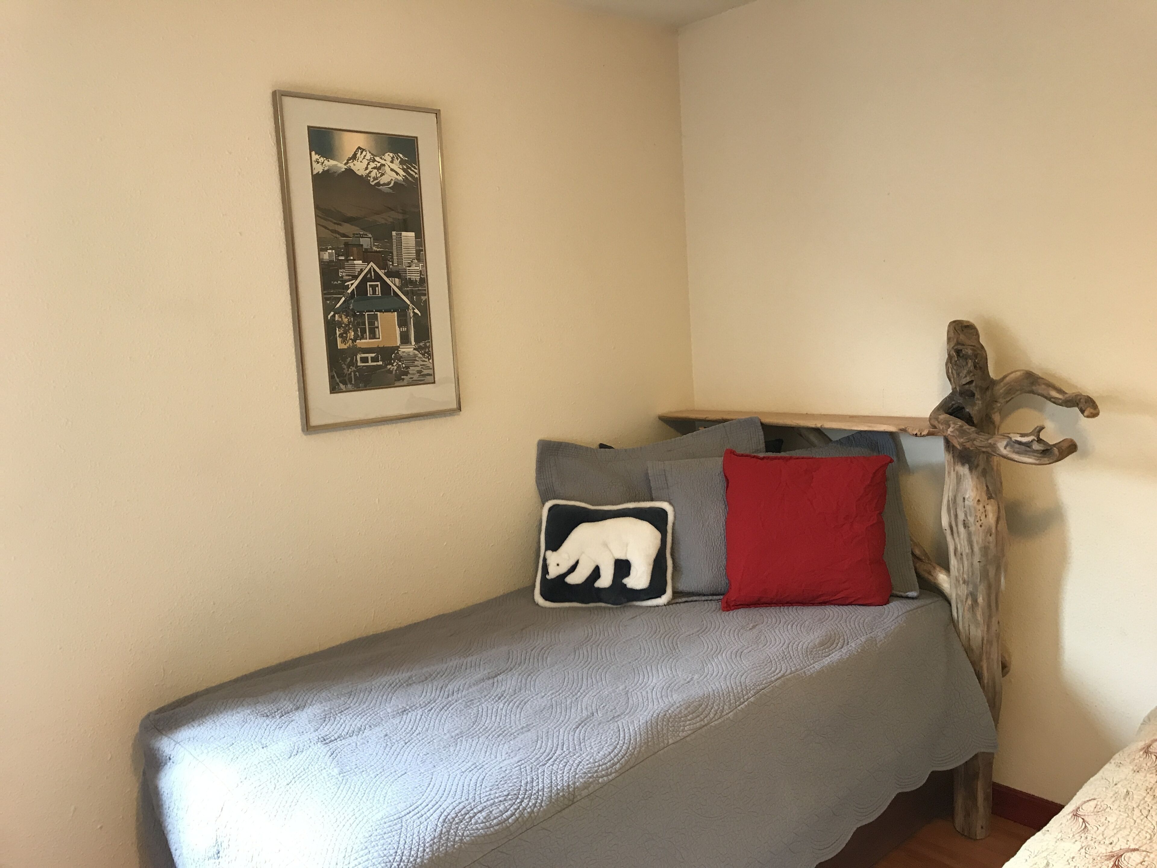 A small, neatly made bed with a gray blanket, a red pillow, and a pillow with a white bear design. A framed picture of a mountain landscape hangs on the wall above the bed.
