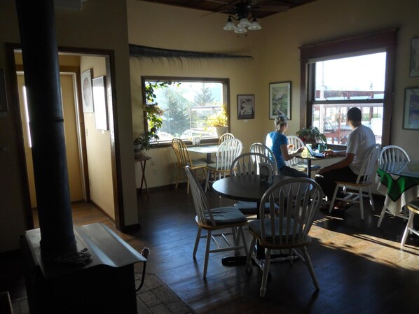 A restaurant with wooden tables and chairs, large windows with views of the outside, and a ceiling fan. - A Swan Nest Inn (Seward)