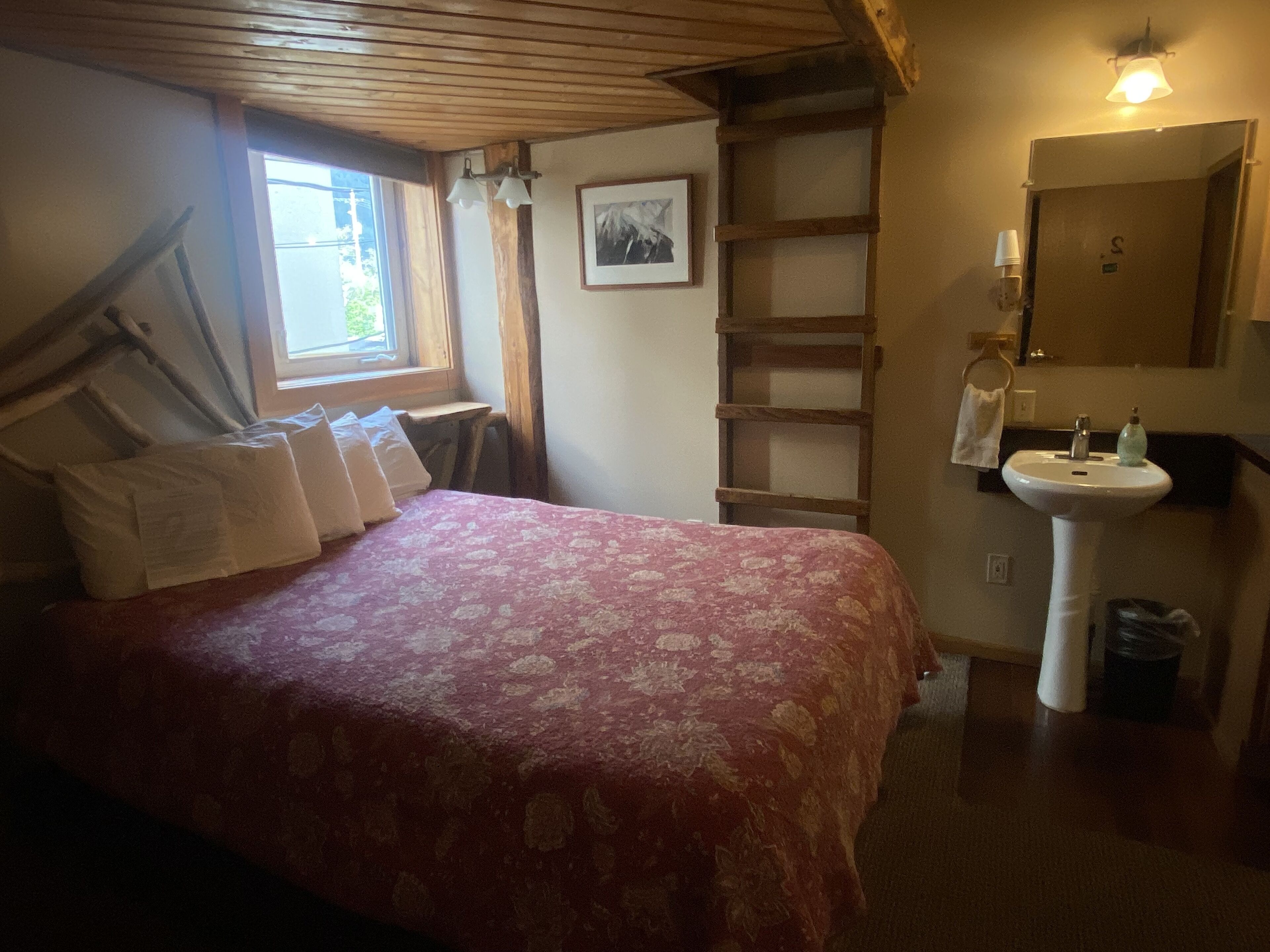 A bedroom with a bed, a window, a ladder, a sink, and a mirror.