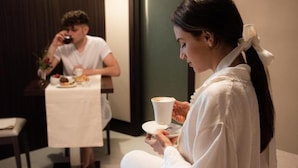Une femme est assise sur un canapé dans une pièce où se trouve une table à manger, et on aperçoit une autre personne à l’arrière-plan.