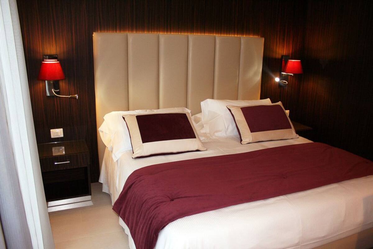 Una habitación de hotel con una cama grande, dos mesitas de noche y apliques rojos en la pared.