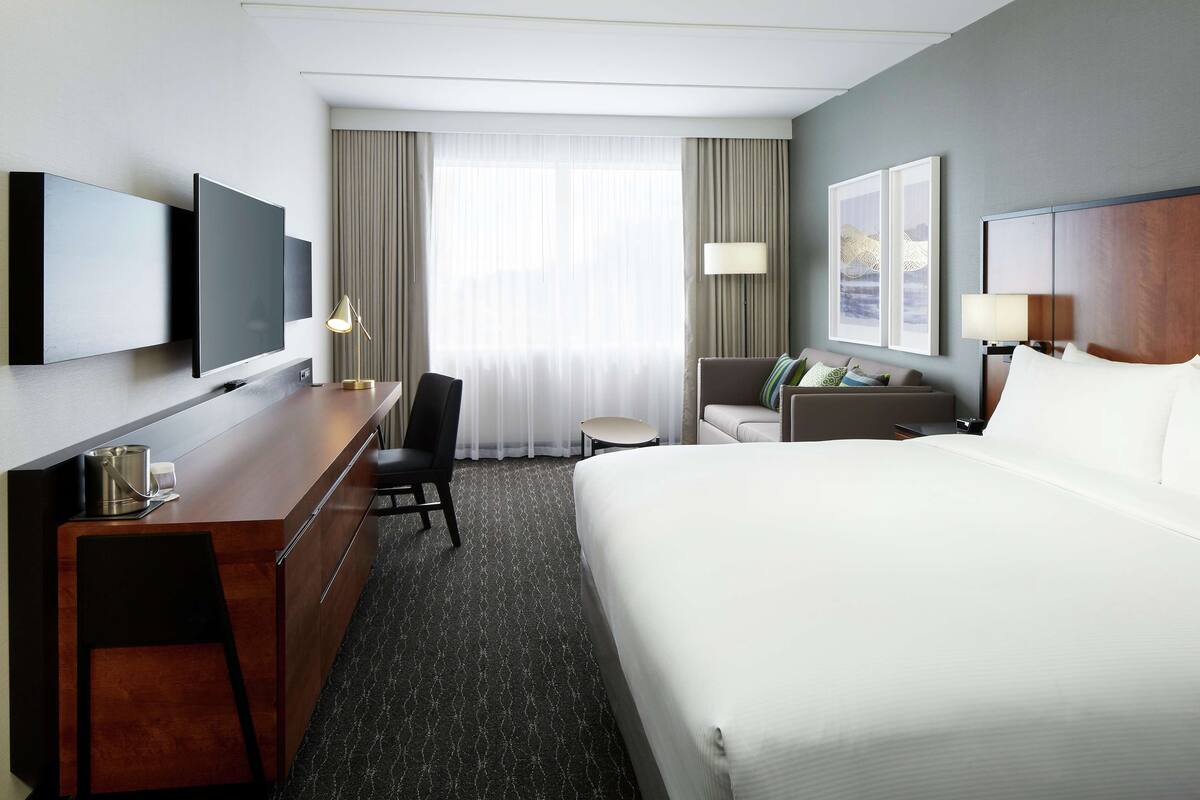 A hotel room with a large bed, a desk with a chair, a TV mounted on the wall, and a window with curtains.