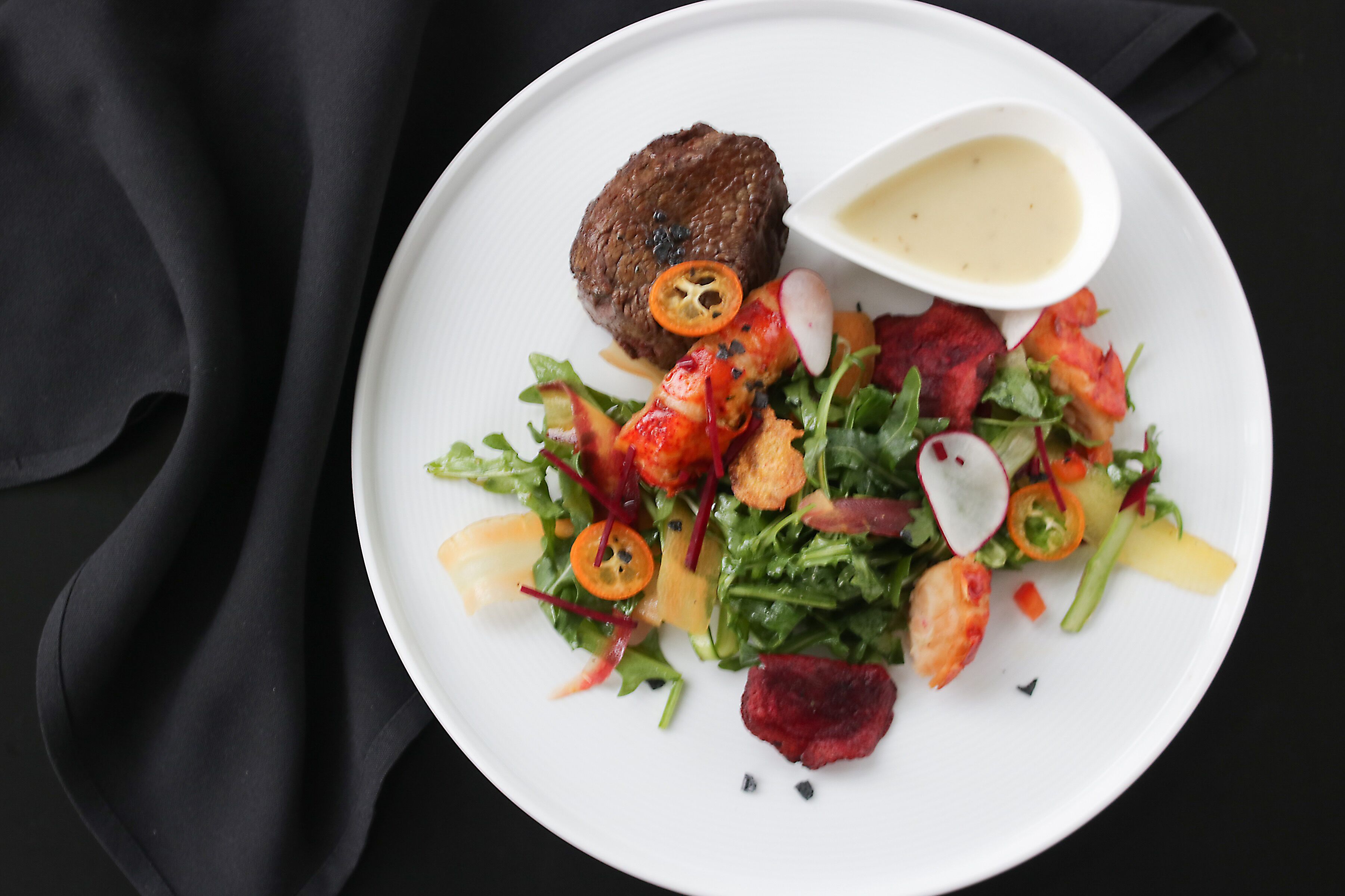 A plate with a salad, a meat patty, and a small bowl of sauce.
