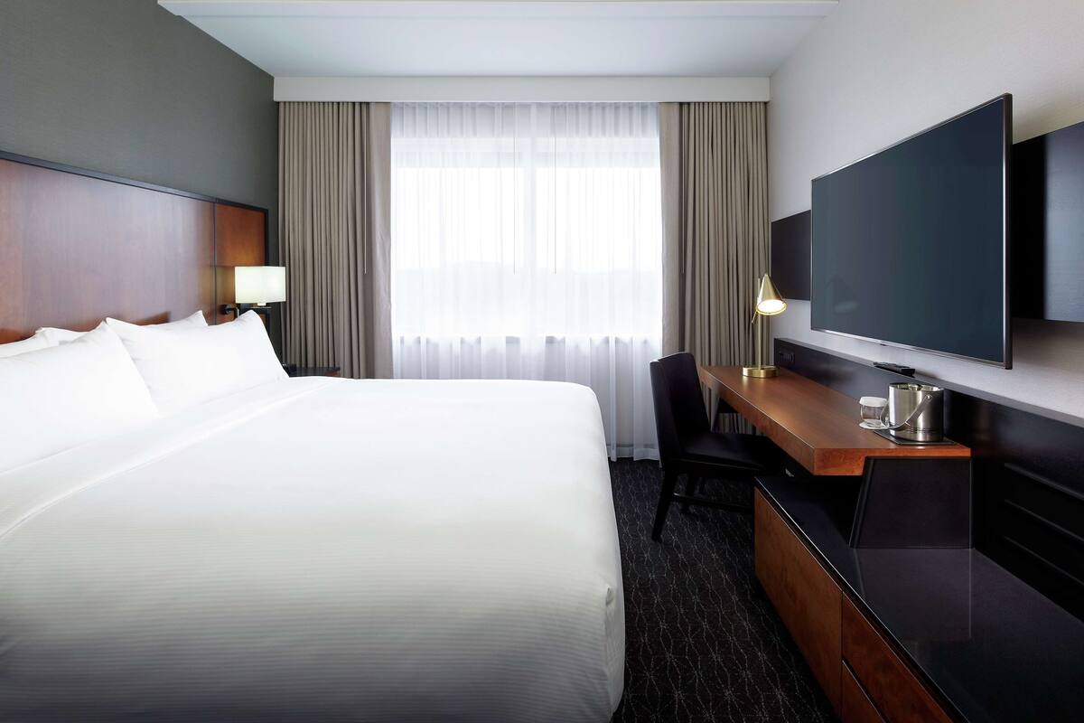 A hotel room with a large bed, a desk with a chair, a flat-screen TV, and a window with curtains.