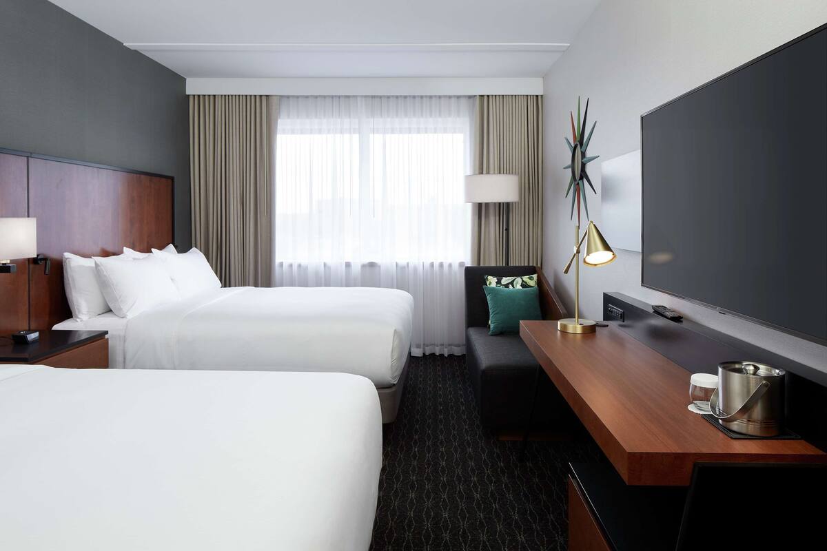 A hotel room with two beds, a desk with a lamp, a TV, and a large window with curtains.