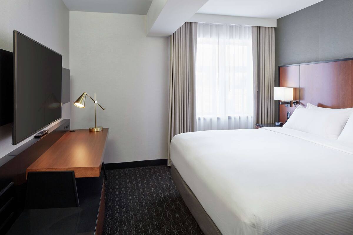 A hotel room with a large bed, a flat-screen TV, a desk with a lamp, and a window with curtains.
