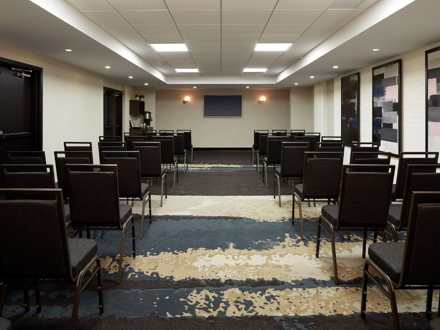Un salón de conferencias con filas de sillas, una pantalla para proyector y piso alfombrado.