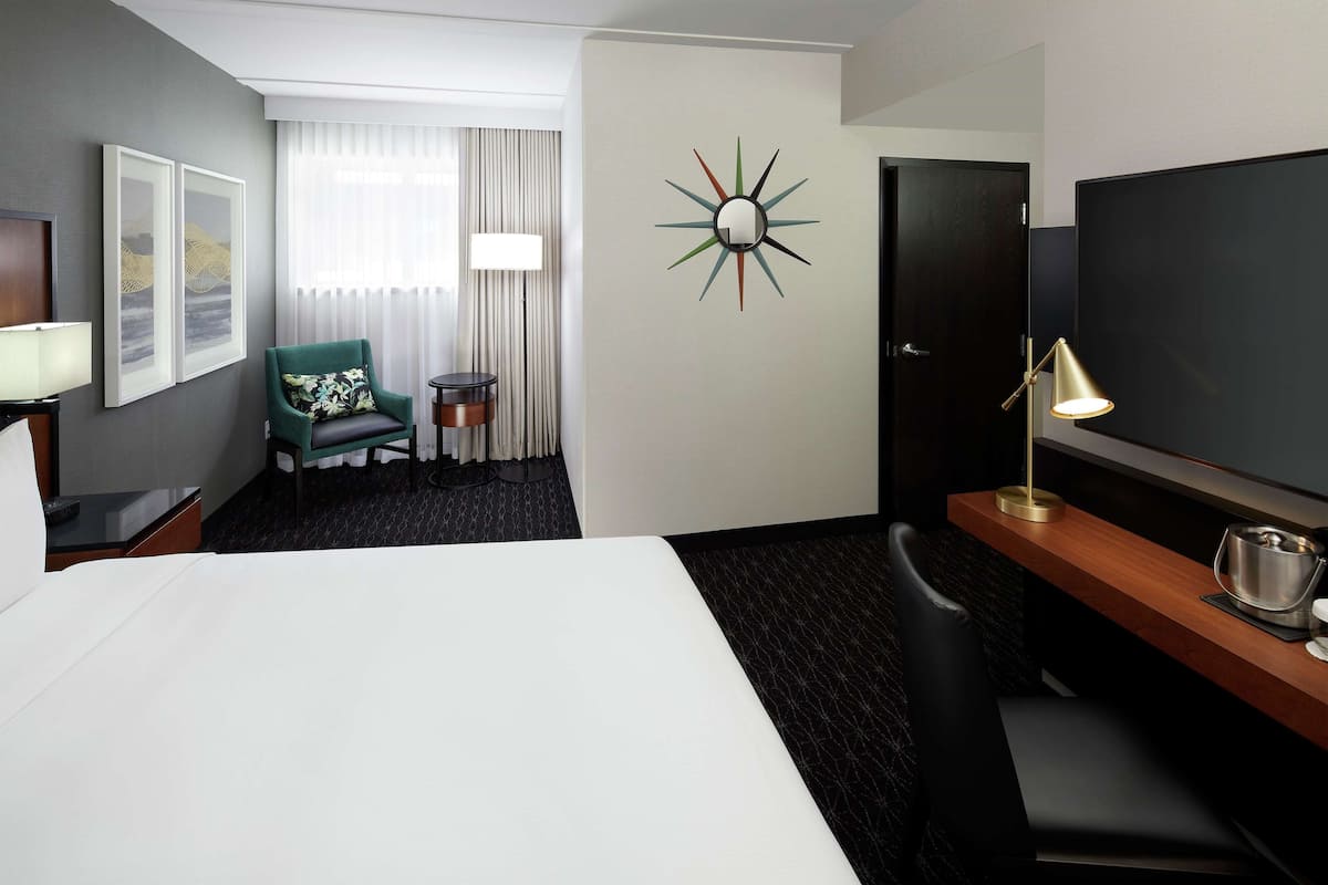 A hotel room with a large bed, a desk with a lamp, a green chair, a TV, and a wall clock.
