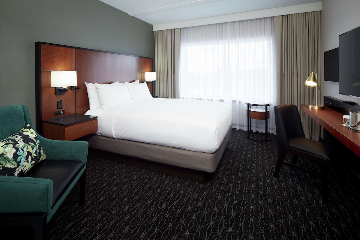 A hotel room with a large bed, two bedside lamps, a desk with a chair, a television, and a view of the outdoors through a window with curtains.