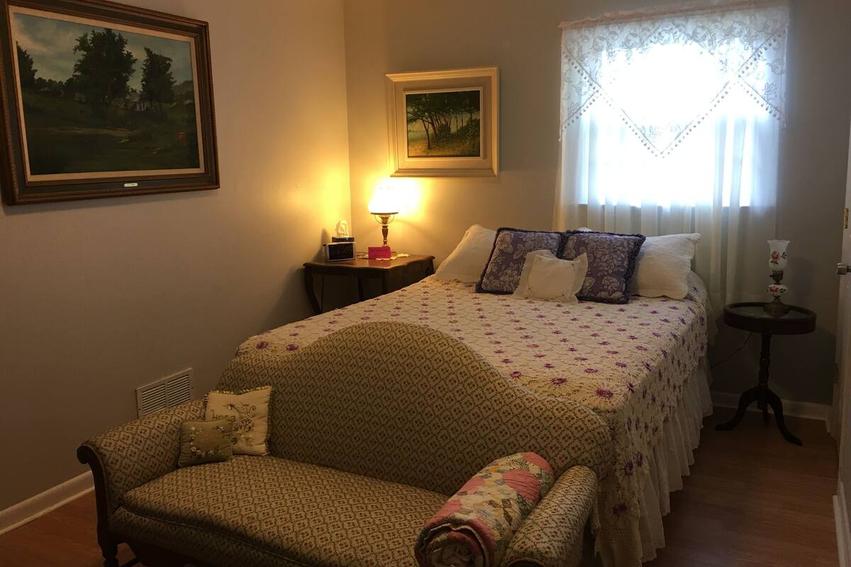 Une chambre confortable avec un lit, une chaise, une table de chevet, une lampe et deux tableaux accrochés au mur.