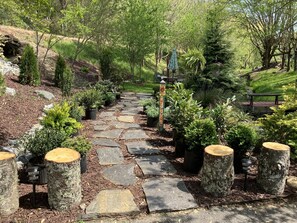 Un chemin de pierres qui traverse un jardin agrémenté d’une pancarte « Bienvenue », entouré de différentes plantes et arbres.