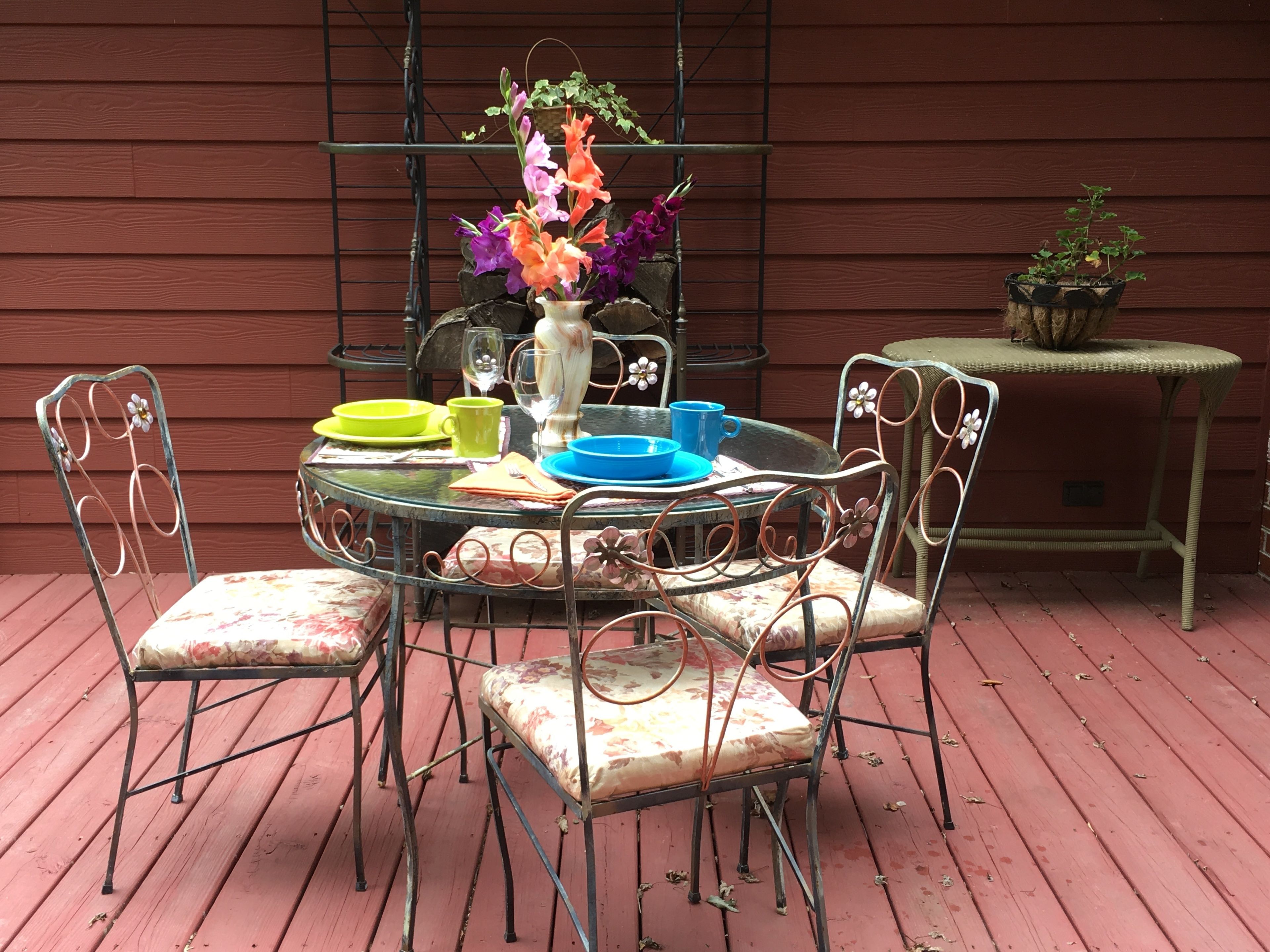 A round metal table with floral cushions, surrounded by metal chairs. A vase with flowers and a small potted plant are on the table.