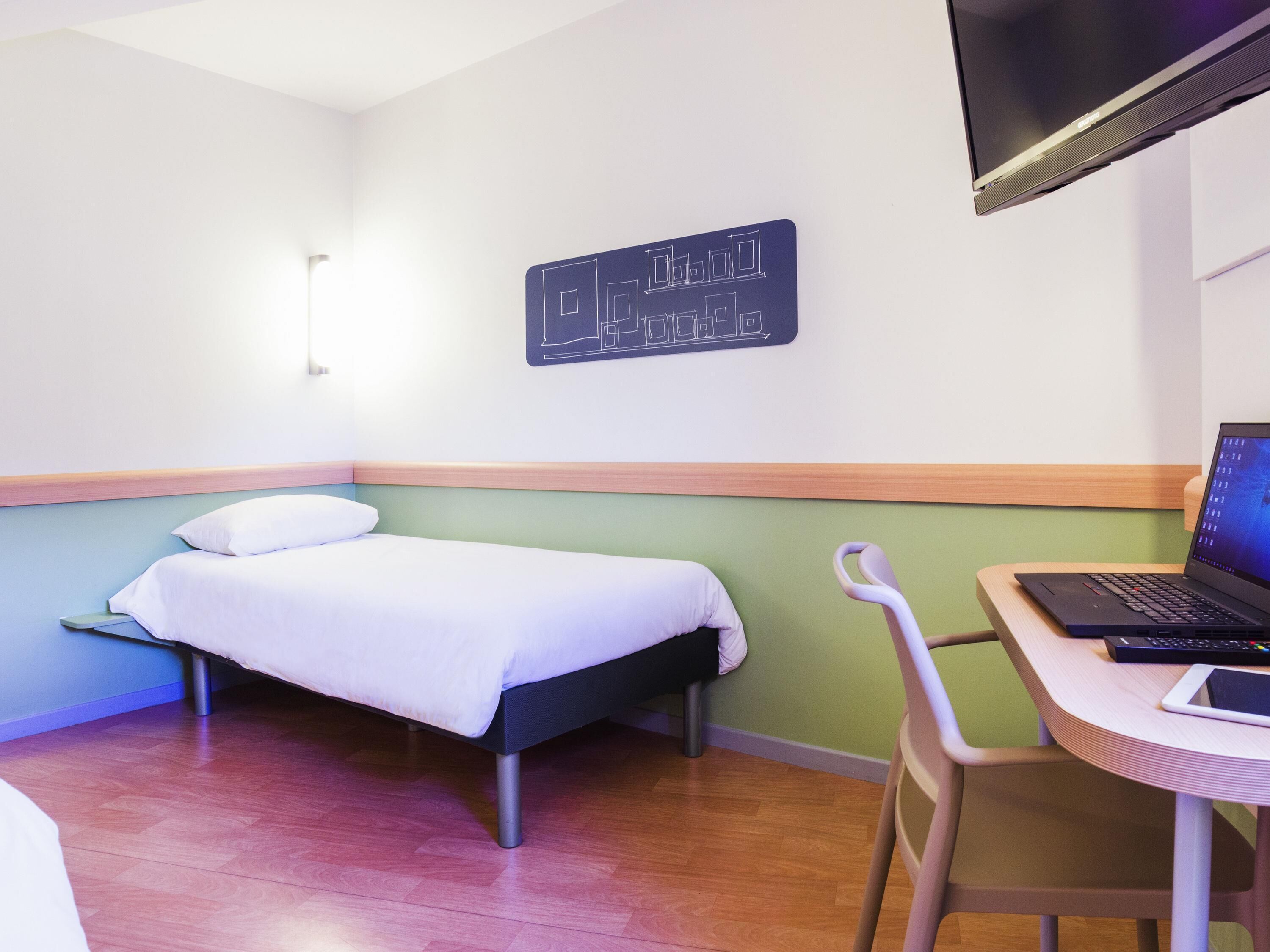 Una habitación de hotel con cama, escritorio con computadora portátil y televisor montado en la pared.