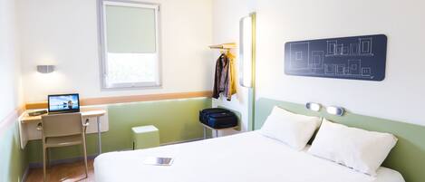 A hotel room with a bed, a desk with a laptop, a suitcase, and a coat hanging on the wall.