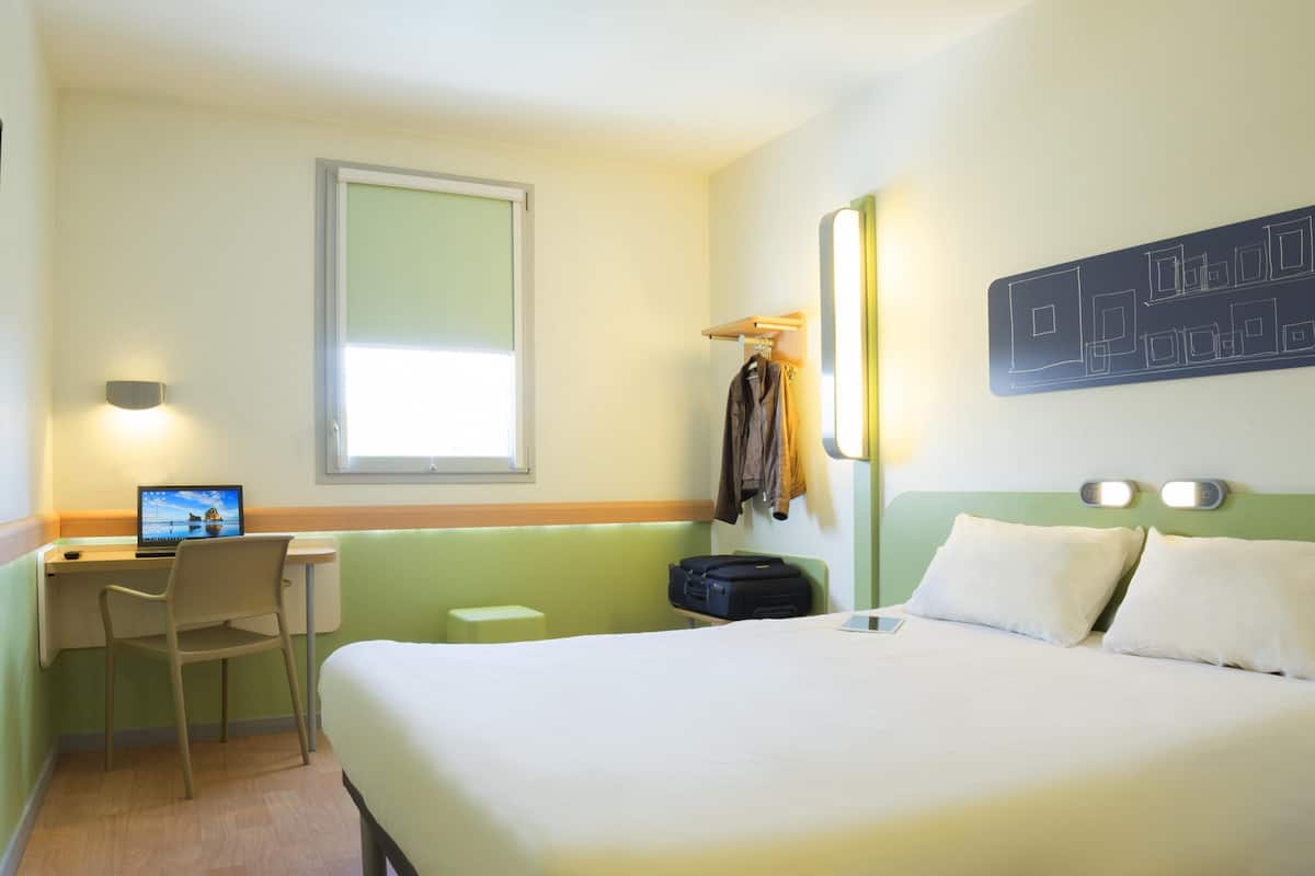 A hotel room with a bed, a desk with a laptop, a chair, a suitcase, and a coat rack.