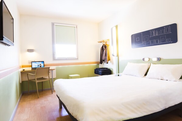 Une chambre d’hôtel équipée d’un lit, d’un bureau avec un ordinateur portable, d’une chaise, d’un porte-manteau et d’un téléviseur fixé au mur.