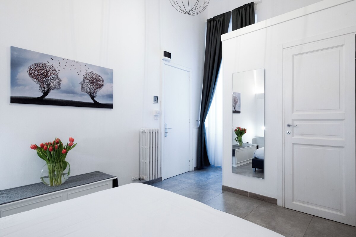 Una habitación de hotel moderna con una cama grande, una mesita de noche con un jarrón de tulipanes rojos, un radiador blanco, una puerta con un espejo y un cuadro de dos árboles con pájaros volando.