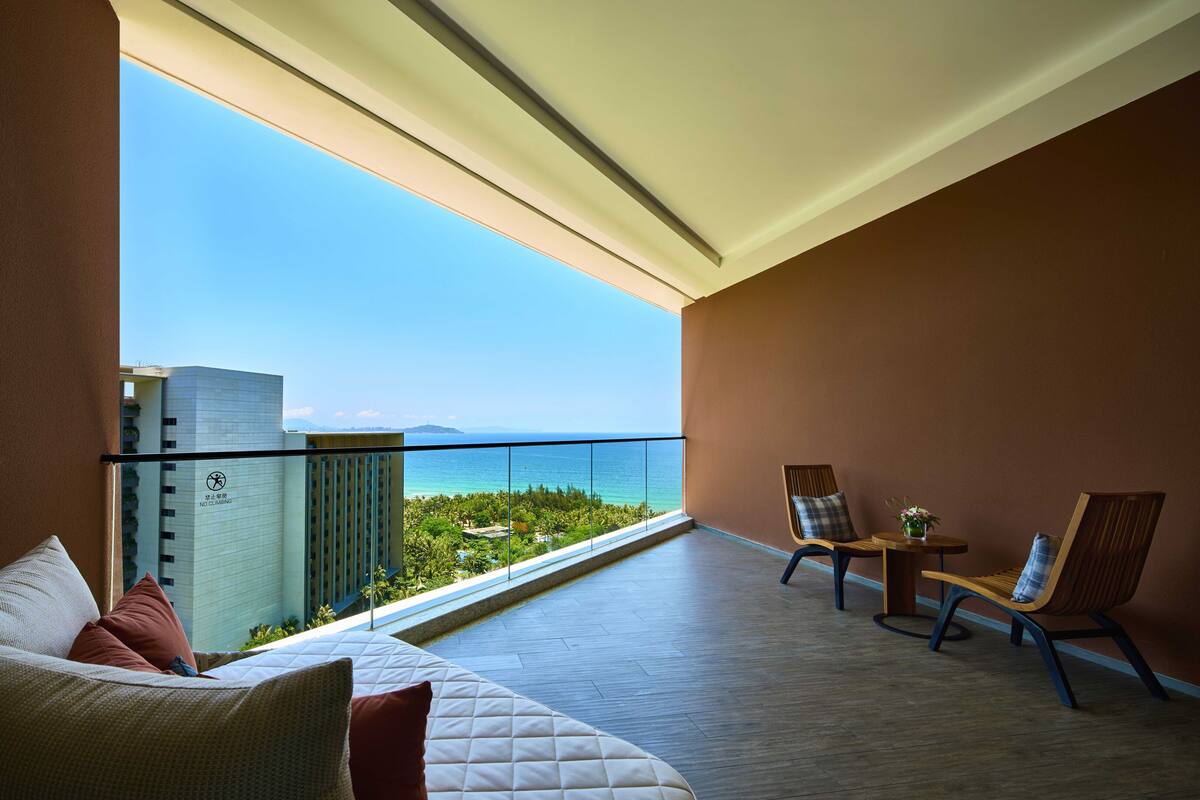 Ein Balkon mit Blick auf das Meer und ein Gebäude. Es gibt ein Bett mit Kissen und einen kleinen Tisch mit einer Vase.