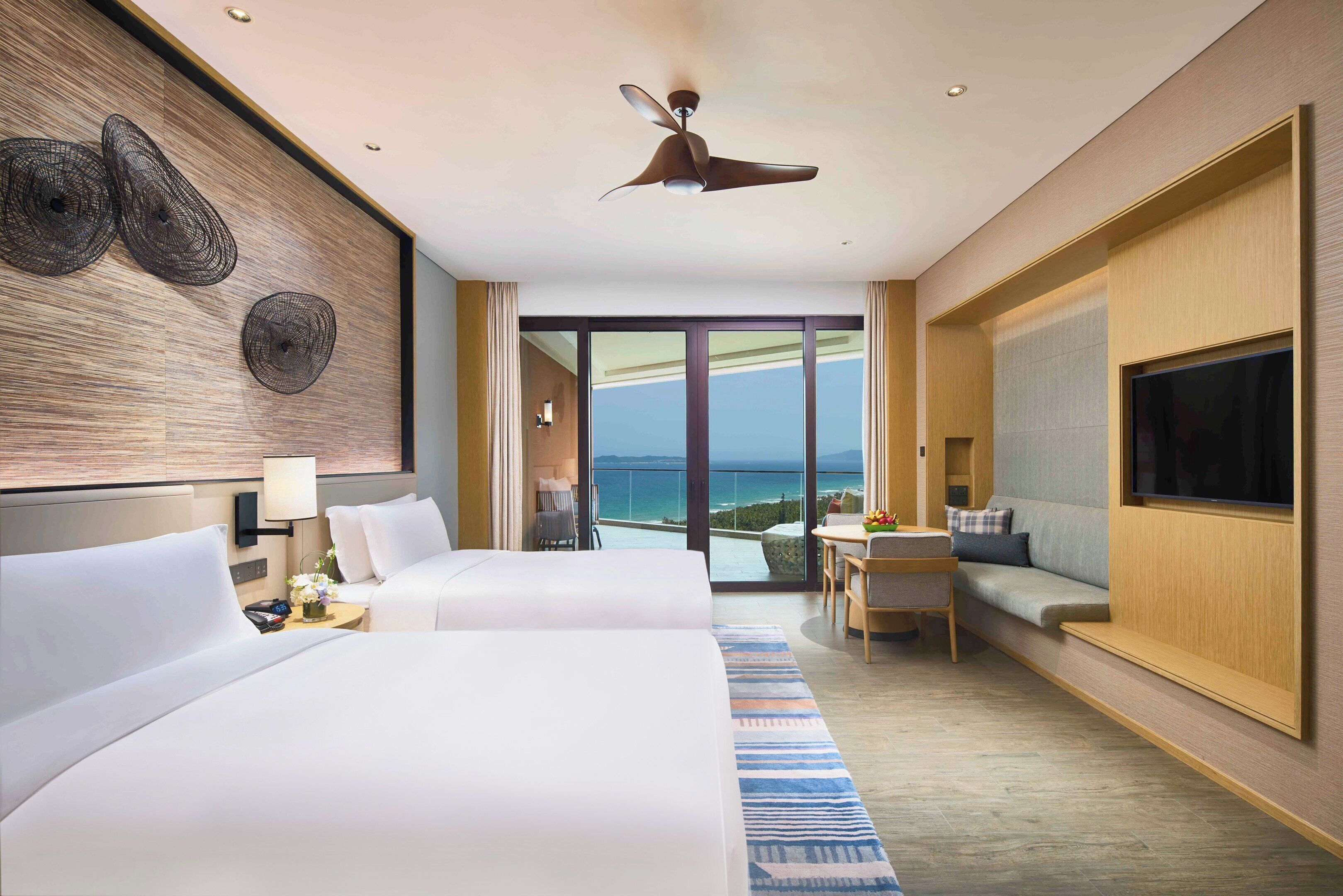 A hotel room with two beds, a TV, a ceiling fan, a large window with a view of the sea, and a balcony with seating.