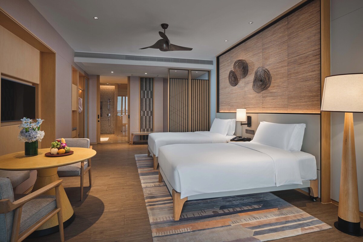 Habitación de hotel con dos camas, zona de comedor con mesa y sillas, zona de ducha y una gran obra de arte en la pared.