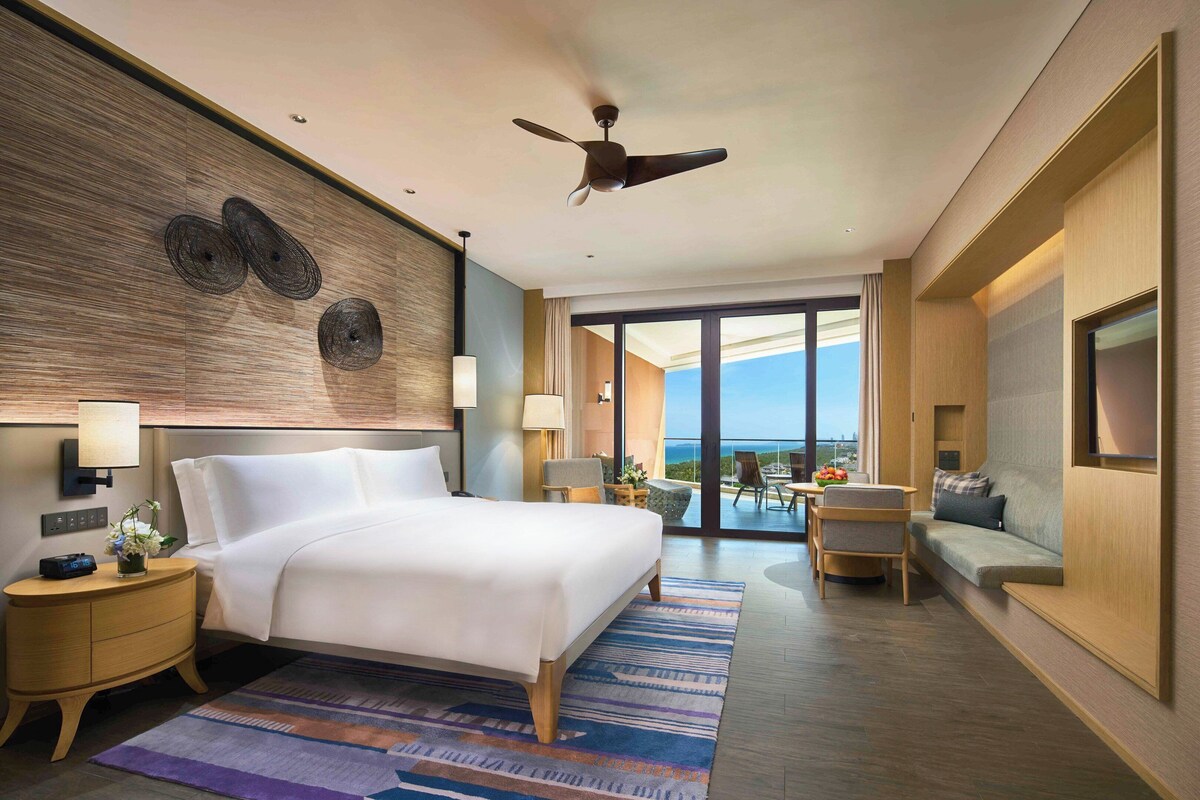 A modern hotel room with a large bed, a ceiling fan, a view of the outdoors, a seating area with a sofa and chairs, and a TV on the wall.