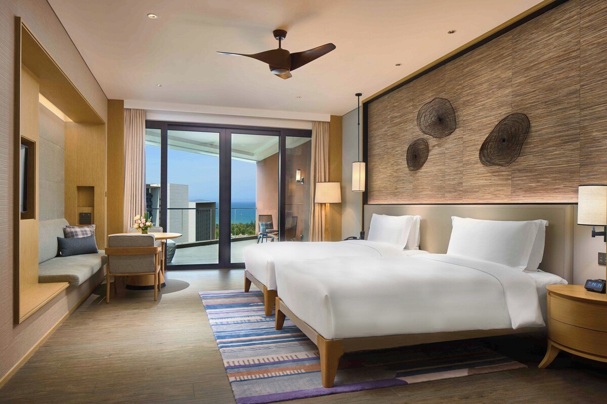 A hotel room with a large bed, a desk, a chair, a balcony with a view, and a ceiling fan.
