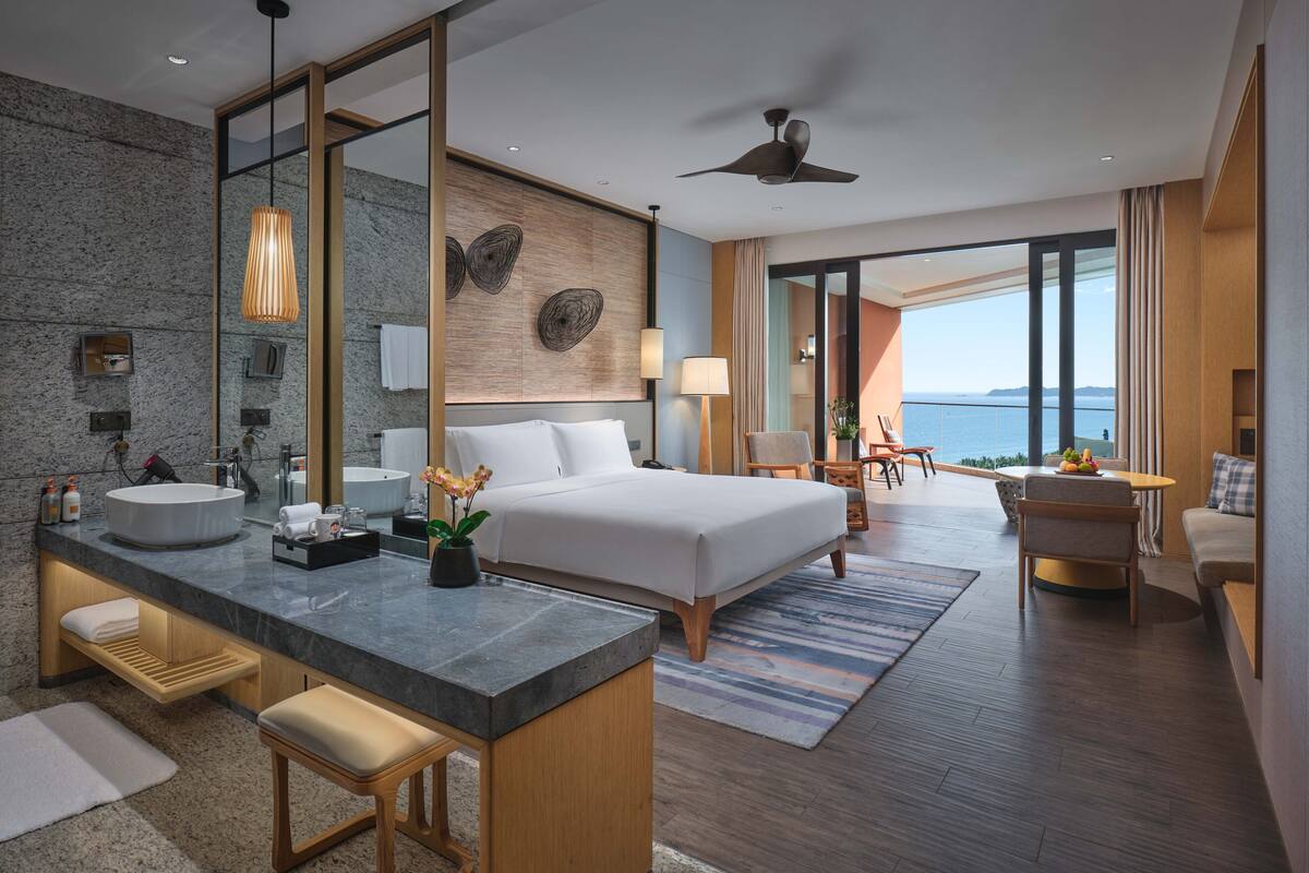 Ein modernes Hotelzimmer mit einem großen Bett, einer Sitzecke mit Blick, einem Badezimmer mit großem Spiegel und einem Balkon mit Aussicht.