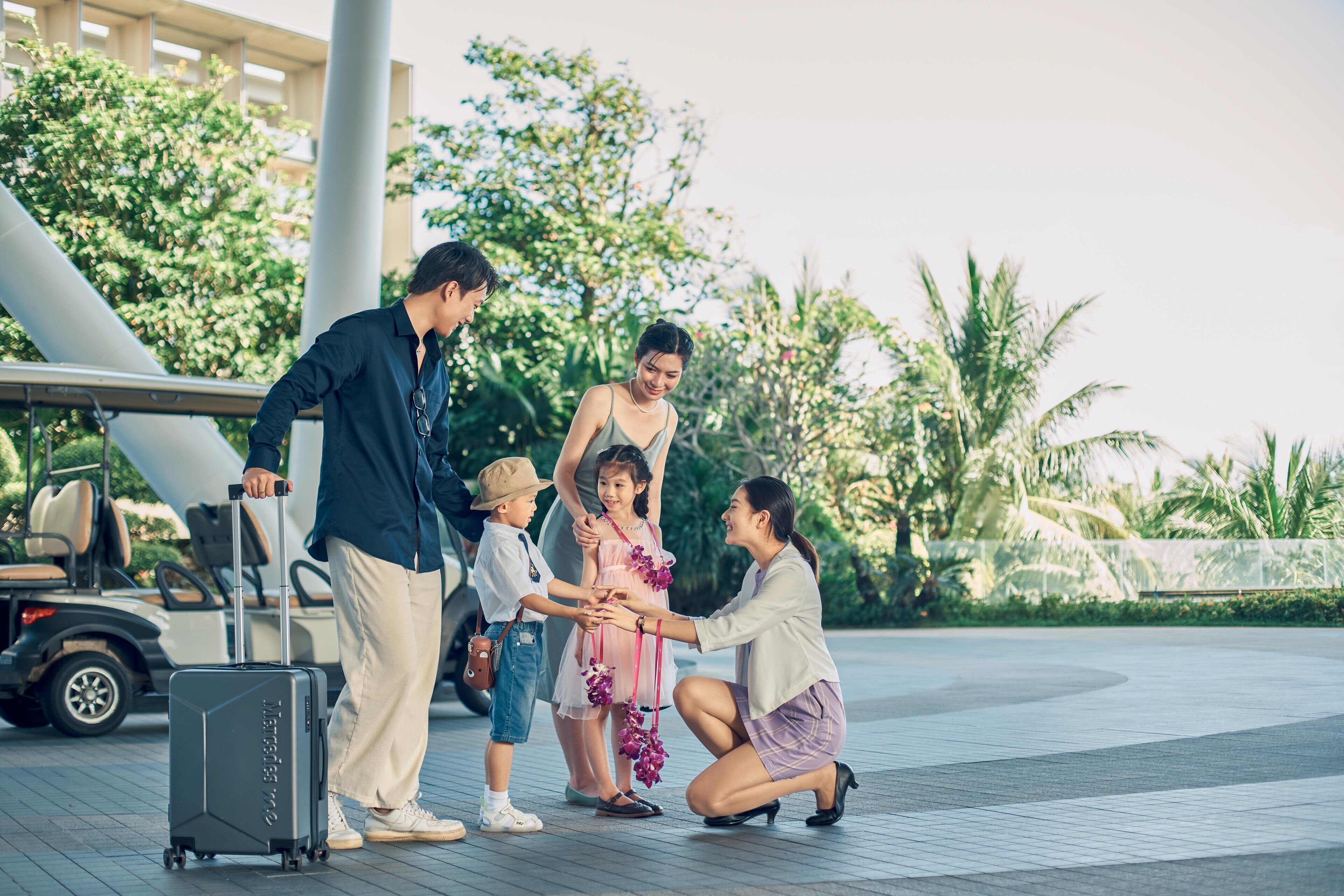 A family with a child and a woman kneeling, standing near a golf cart and a suitcase.