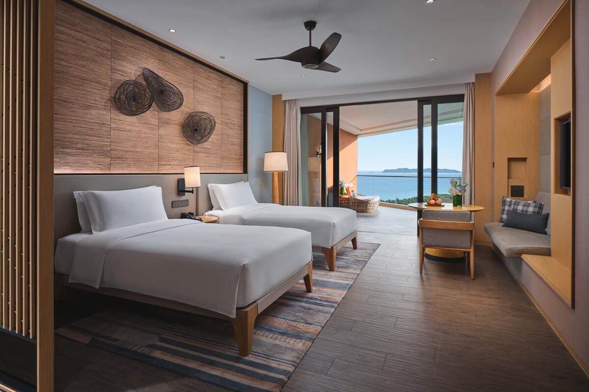 A modern hotel room with two beds, a balcony with a view, a ceiling fan, and a small seating area with a table.
