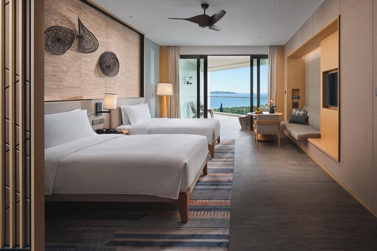Habitación de hotel con dos camas, ventilador de techo, balcón con vistas y televisor en la pared.