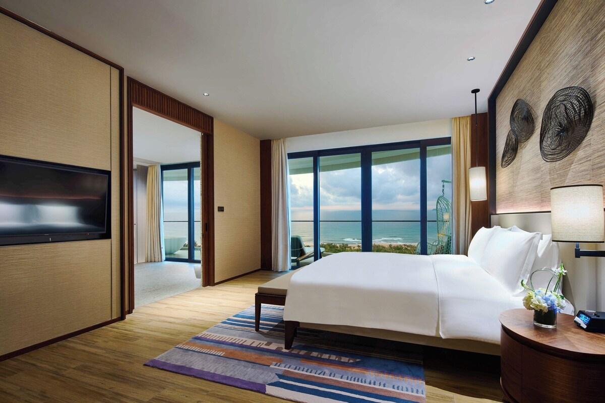 Habitación de hotel con una cama grande, televisión, vista al océano y un ventanal amplio.