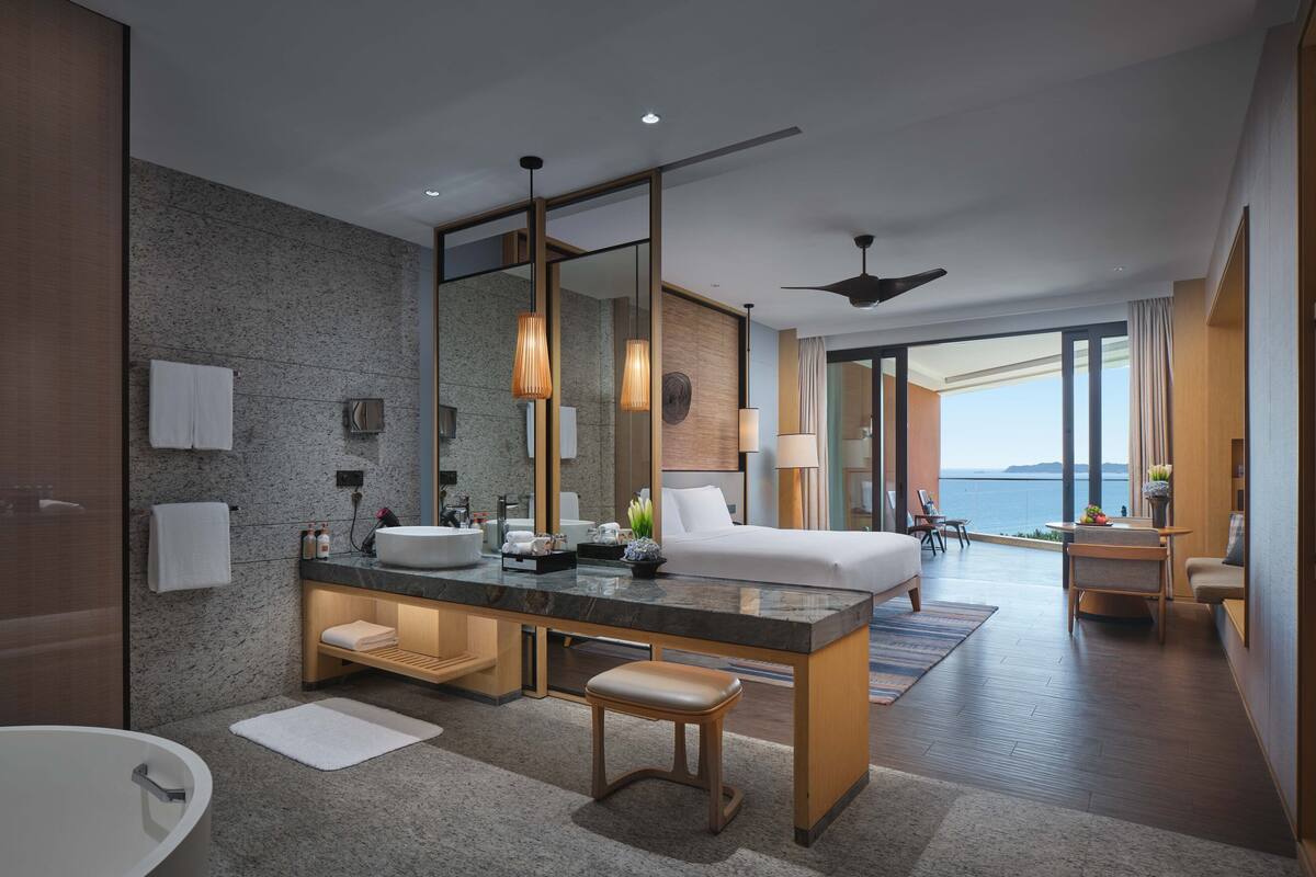 Ein modernes Hotelzimmer mit einem großen Bett, einem separaten Schreibtischbereich, einem Badezimmer mit Badewanne und Meerblick.