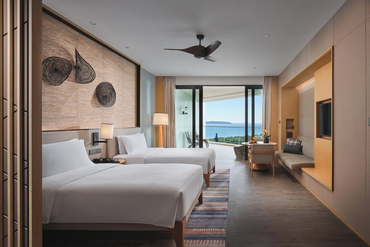 Habitación de hotel con dos camas, ventilador de techo, balcón con vistas y televisor en la pared.