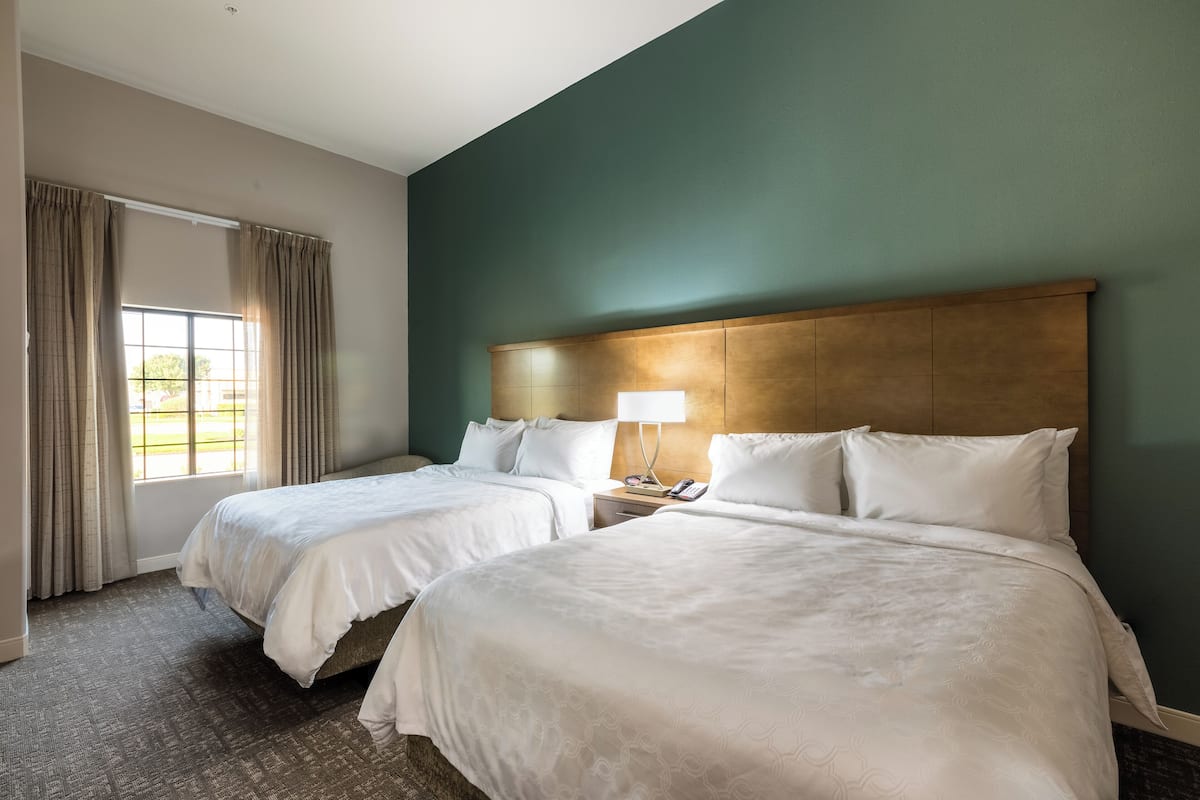 A hotel room with two beds, a wooden headboard, a nightstand with a lamp, and a window with curtains.