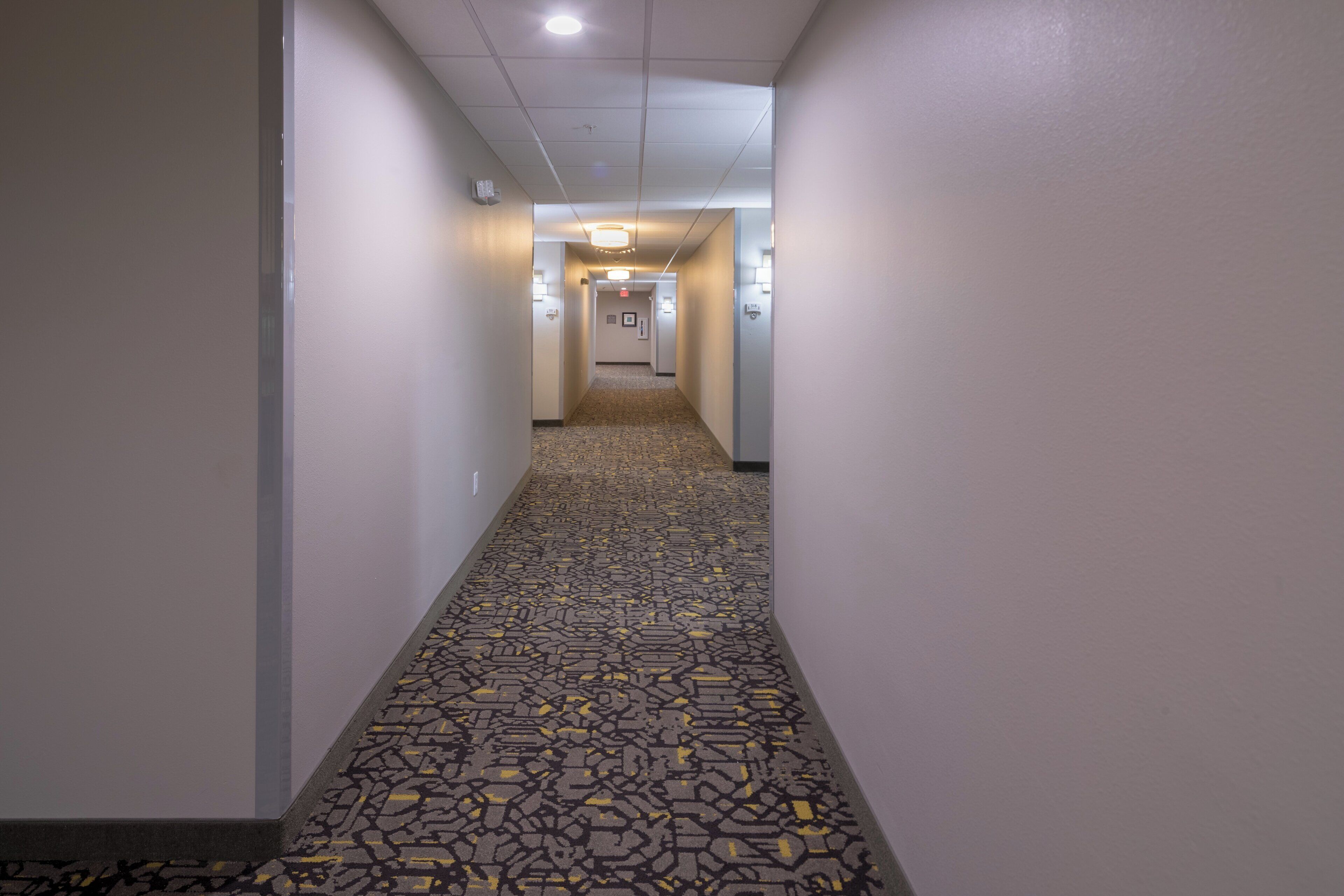 Un pasillo de hotel con alfombra estampada, paredes blancas e iluminación empotrada en el techo.