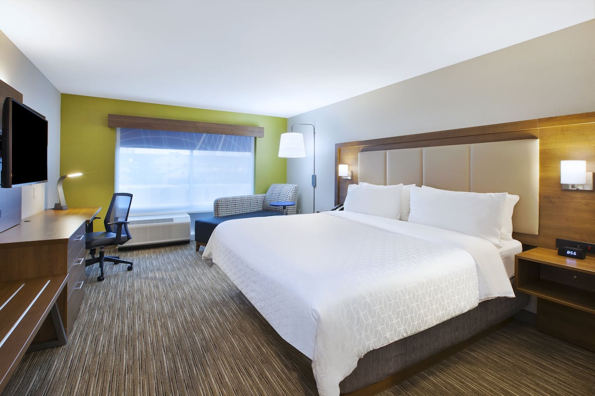 Une chambre d’hôtel avec un grand lit, un bureau avec une chaise, une télévision et une fenêtre avec des stores.