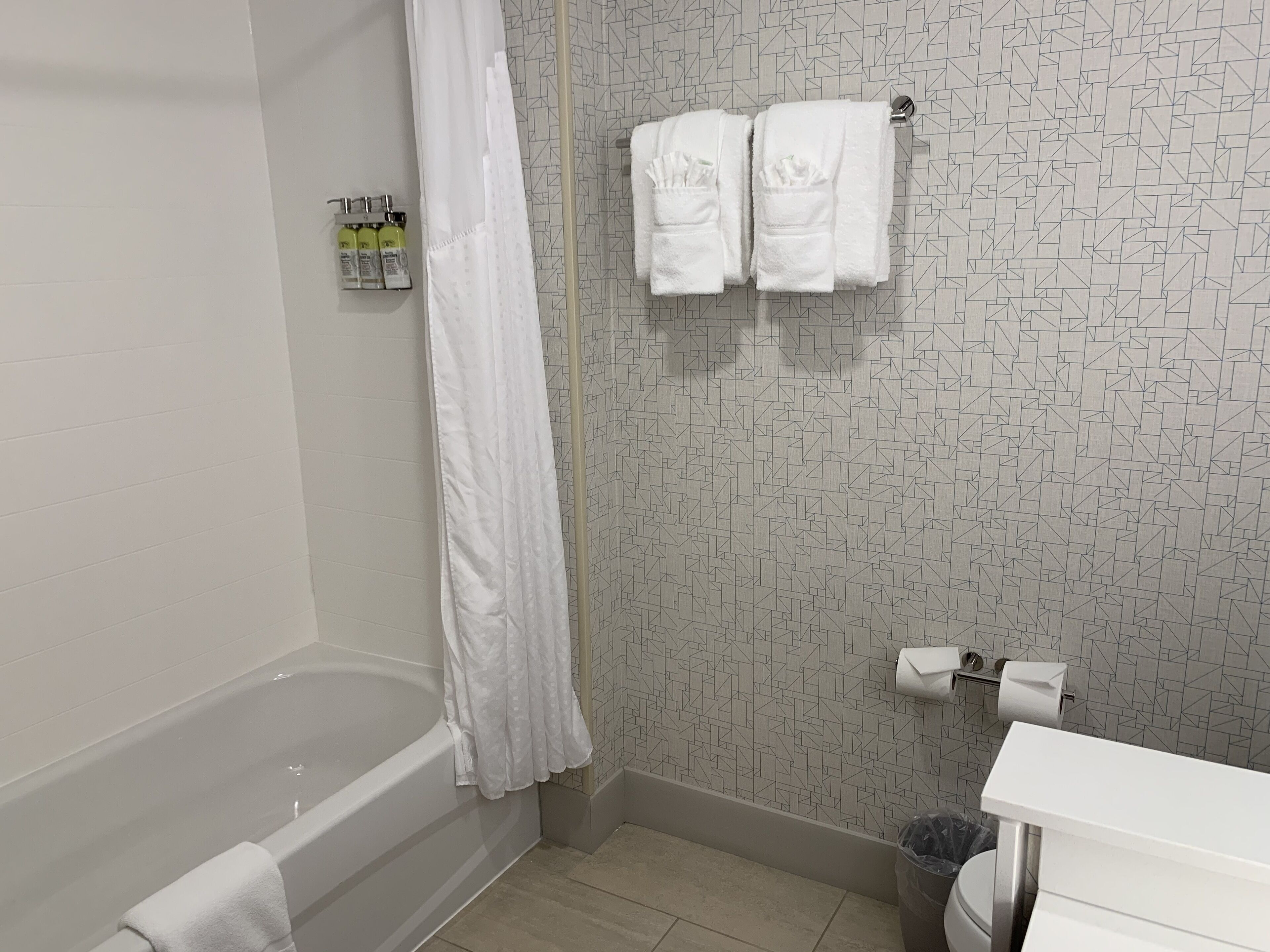 Un baño con bañera, cortina de ducha, toallero con dos toallas, dispensador de papel higiénico y un cubo de basura.