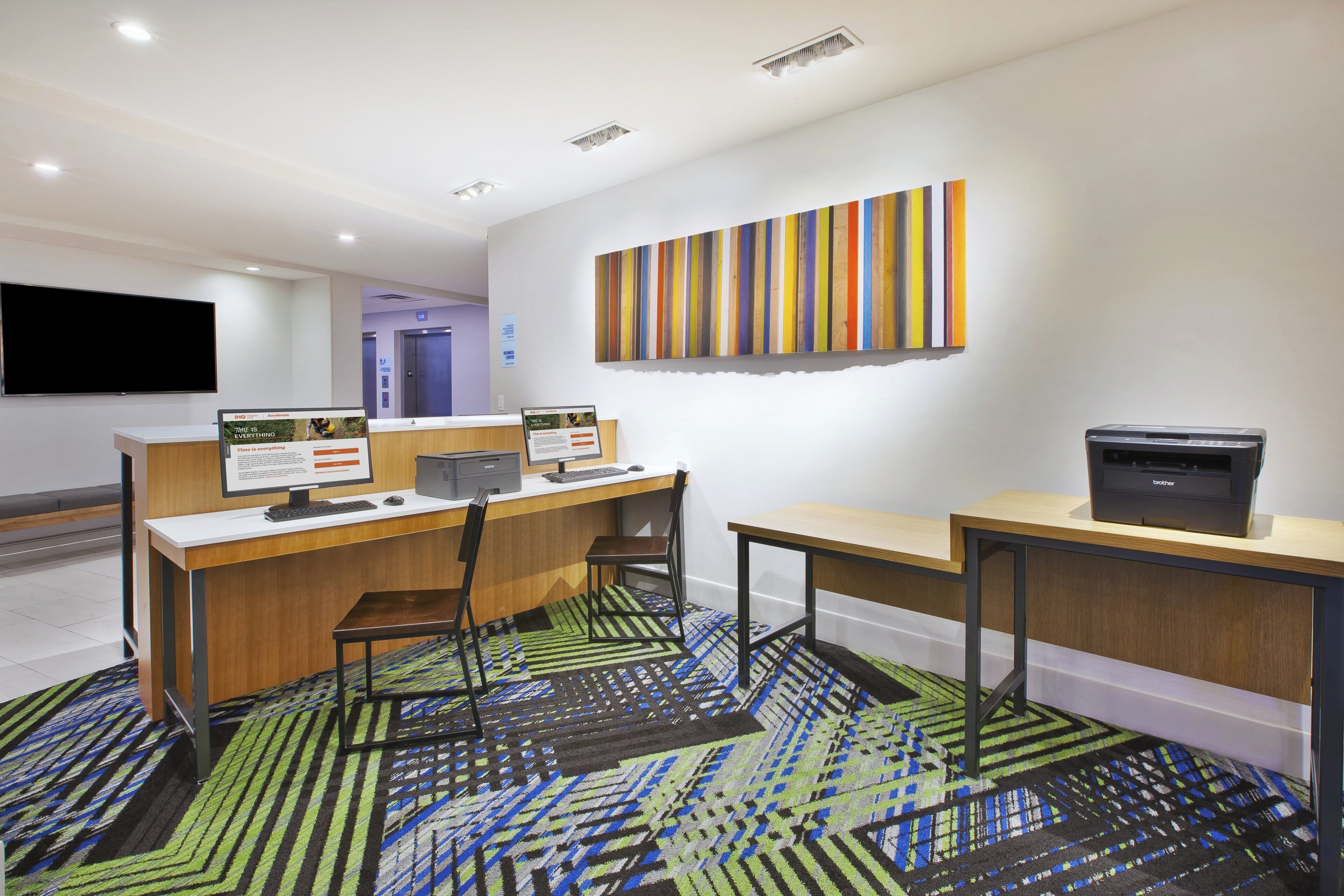 Un espacio de oficina moderno con un mostrador de recepción, dos puestos de trabajo con computadoras, una impresora y un colorido mural a rayas.