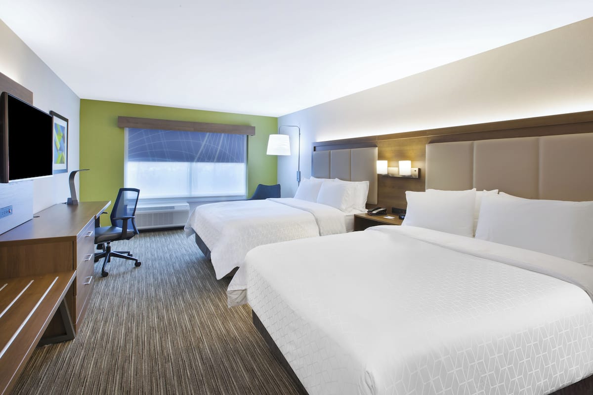 Une chambre d’hôtel avec deux lits, un bureau, une chaise, une télévision et une fenêtre avec des stores.