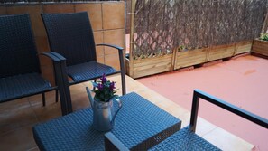 Un patio con dos sillas de mimbre, una mesa pequeña con una jarra de metal y flores, y una jardinera de madera con plantas.