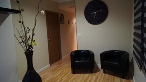 Una habitación con piso de madera, una silla negra, un sillón negro y un reloj de pared.