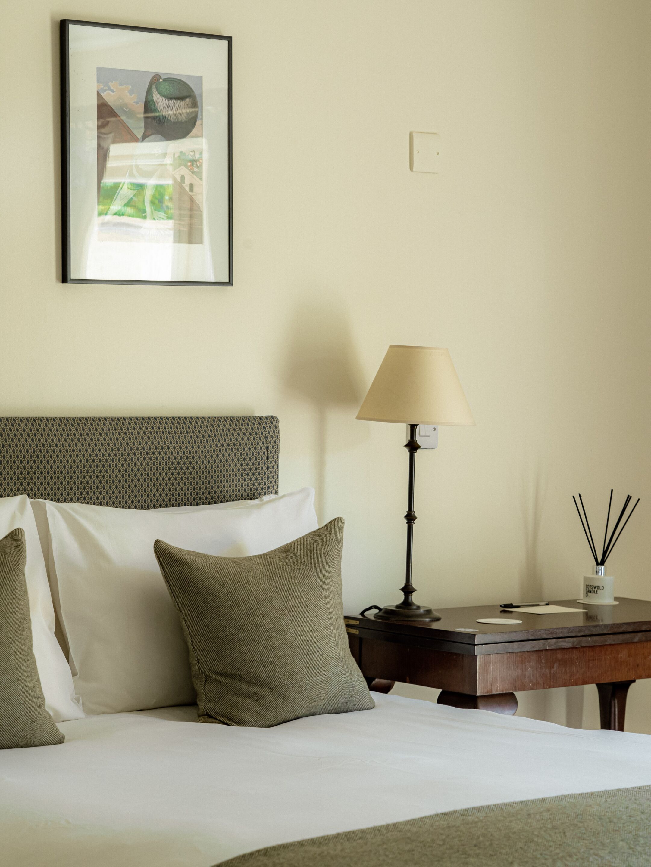 A hotel room with a bed, pillows, a bedside table with a lamp, a framed picture on the wall, and a small table with a candle and reed diffuser.