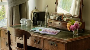 A wooden desk with drawers, a chair, a mirror, a window with curtains, and a vase with flowers. - The Bell Inn (Lechlade)