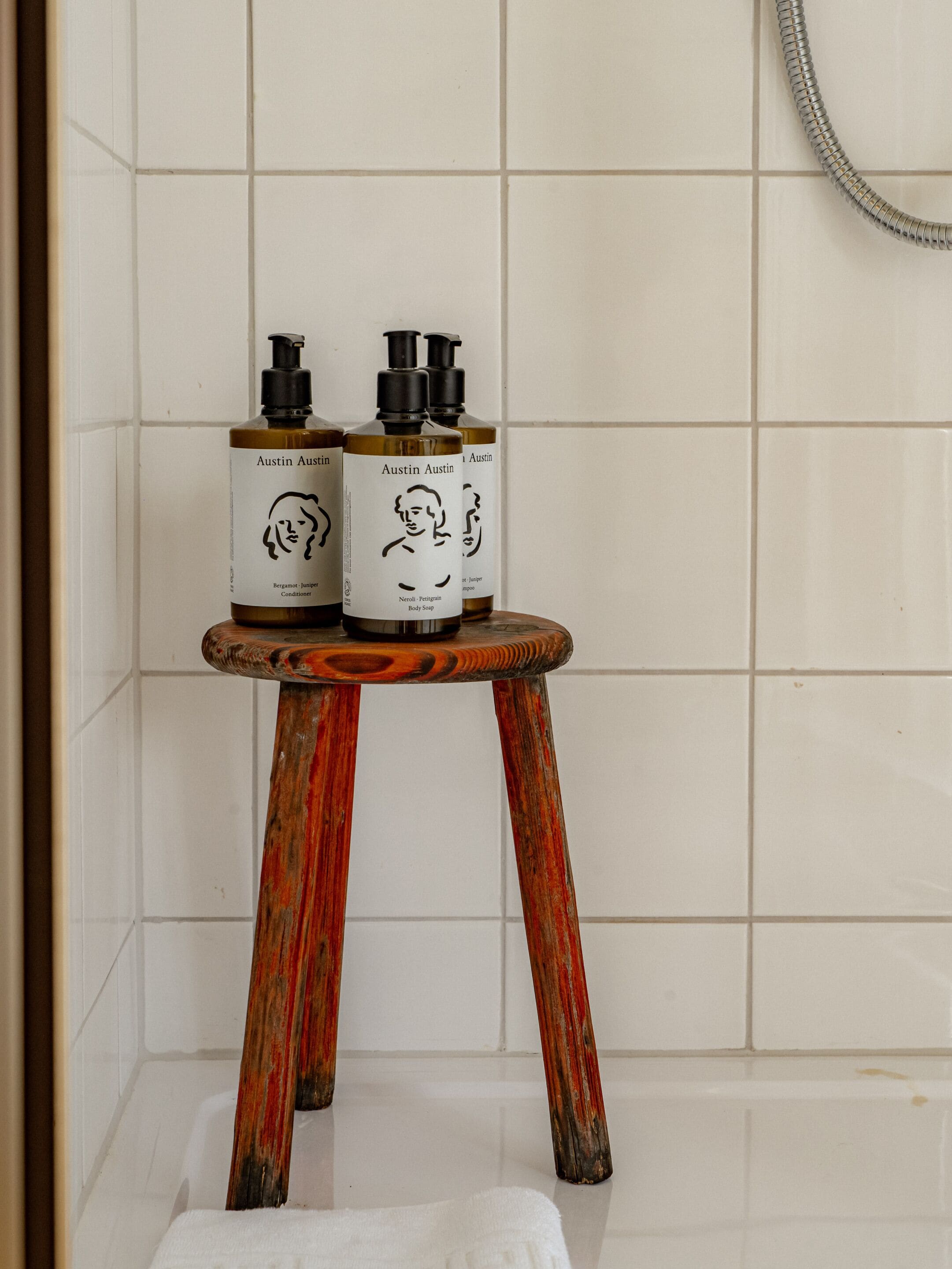 Un baño con paredes de azulejos blancos, un taburete de madera con tres botellas de gel de ducha y una toalla blanca en el suelo.