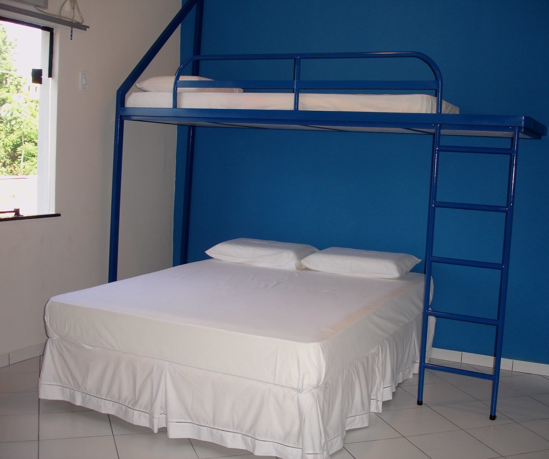 A bunk bed with a white mattress and pillows, a blue metal ladder, and a window with a view of greenery outside.
