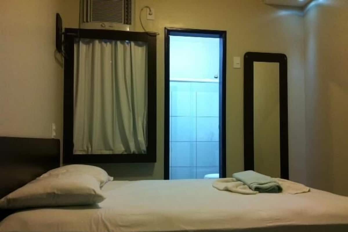 A hotel room with a bed, a window with curtains, a mirror, and a wall-mounted heater.