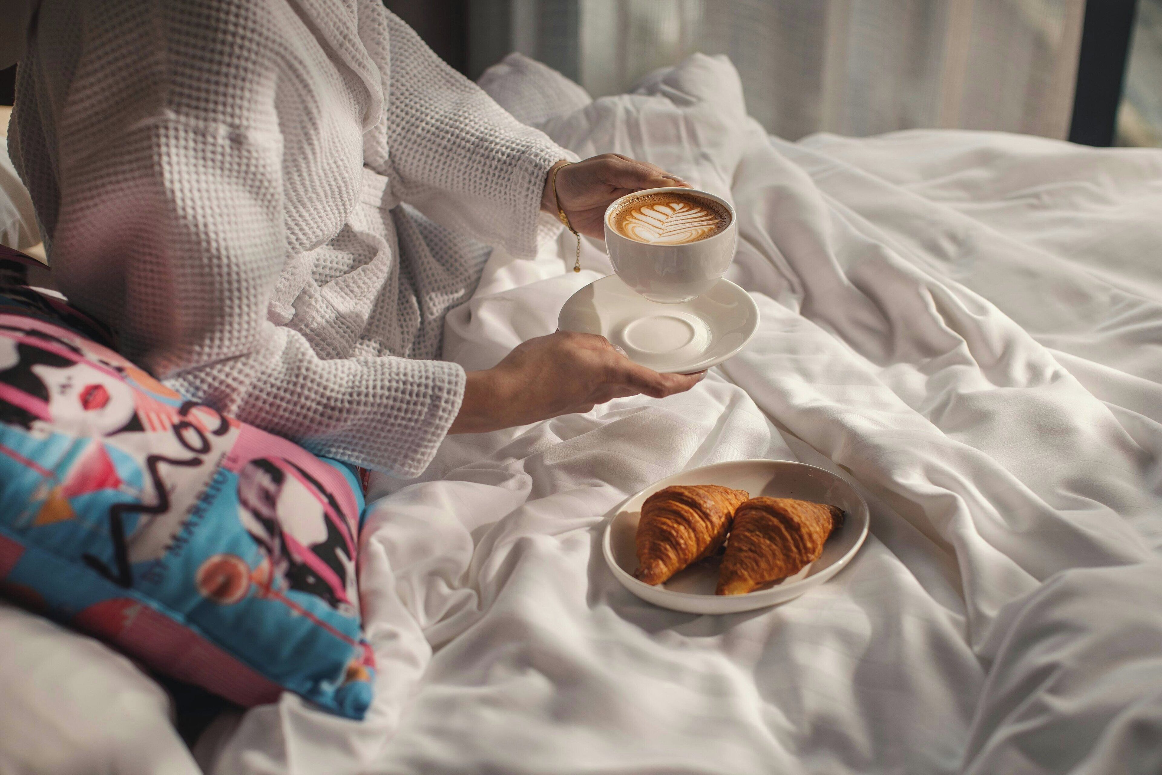 En person holder en kaffekop og en tallerken med croissanter på en seng med hvide sengetæppe.